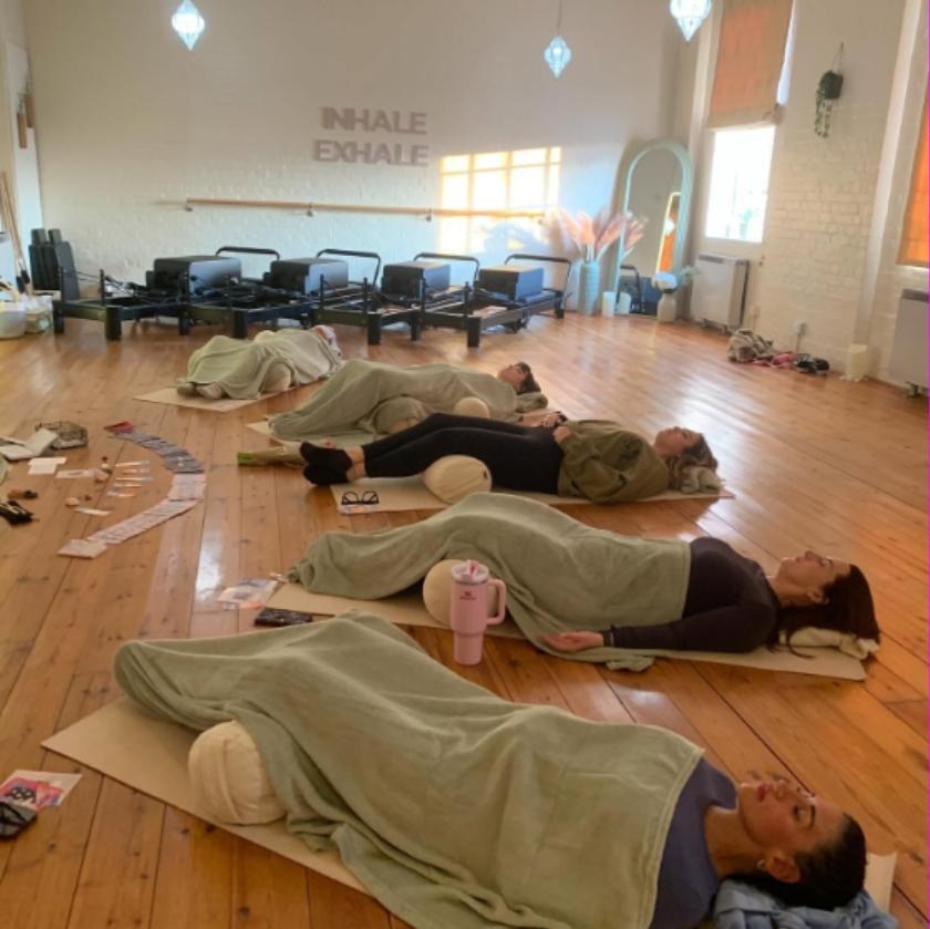 A group Reiki circle in progress — participants resting under sage-green blankets on mats in a light, wooden-floored studio.