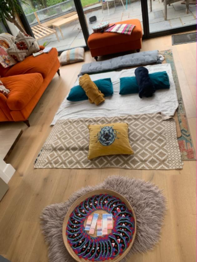 A room laid out for a private group circle: bolsters, cushions and blankets arranged on rugs beside garden doors, with an oracle card spread on a sheepskin.